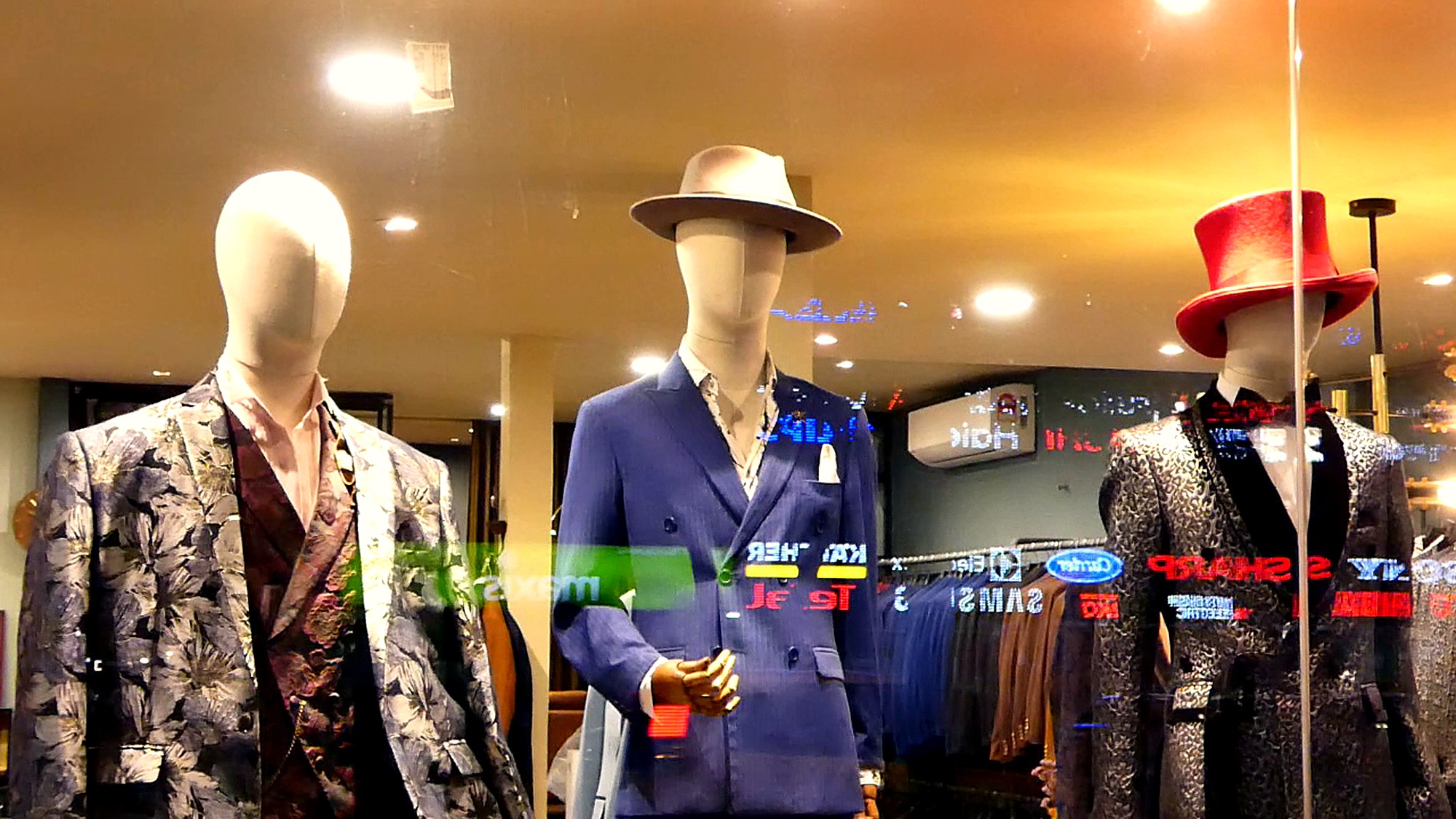 Three mannequins are displayed in a shop window, each wearing a different suit and hat