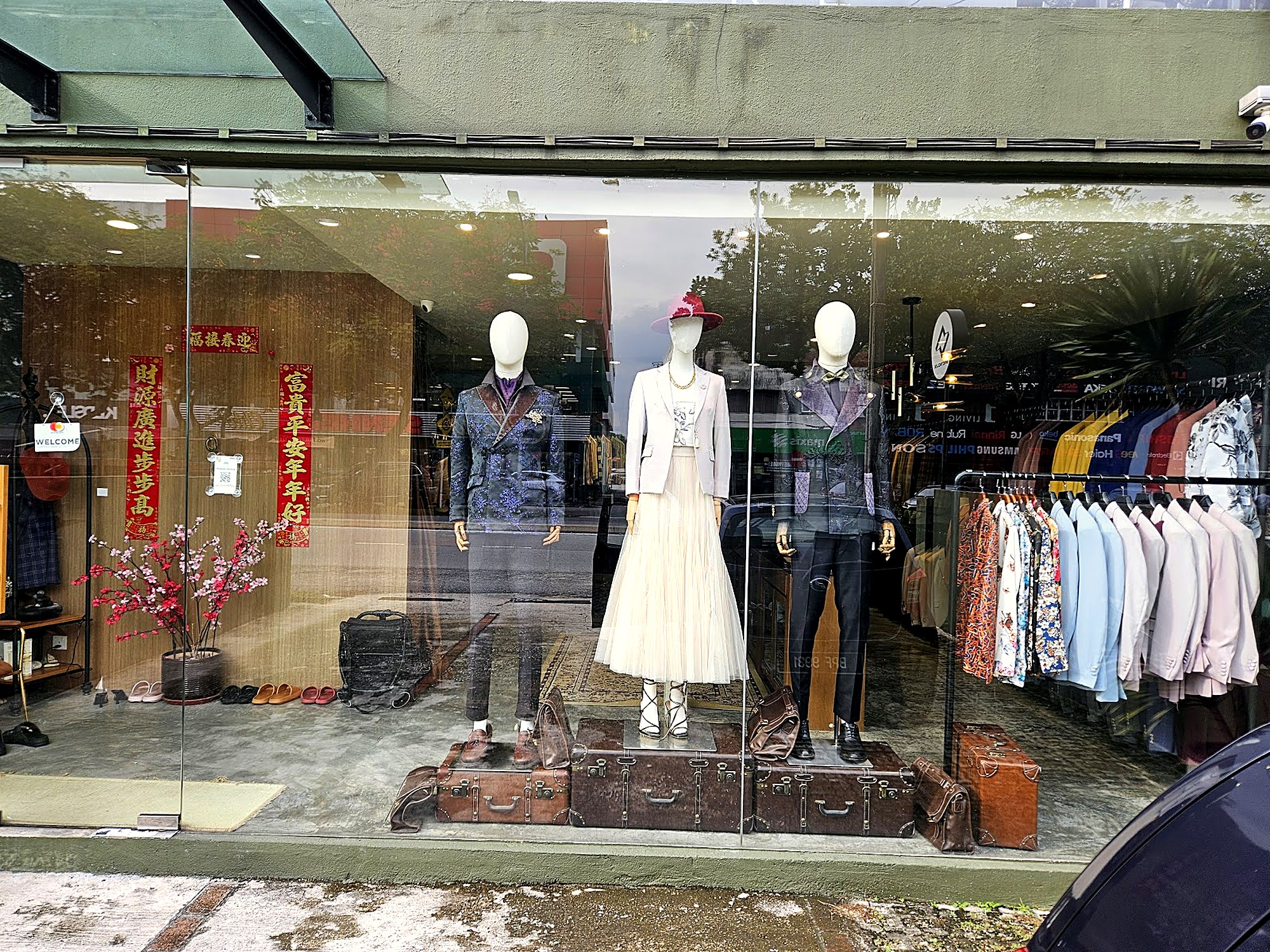 Three mannequins are displayed in a shop window, dressed in suits and a dress, with clothing racks visible behind them