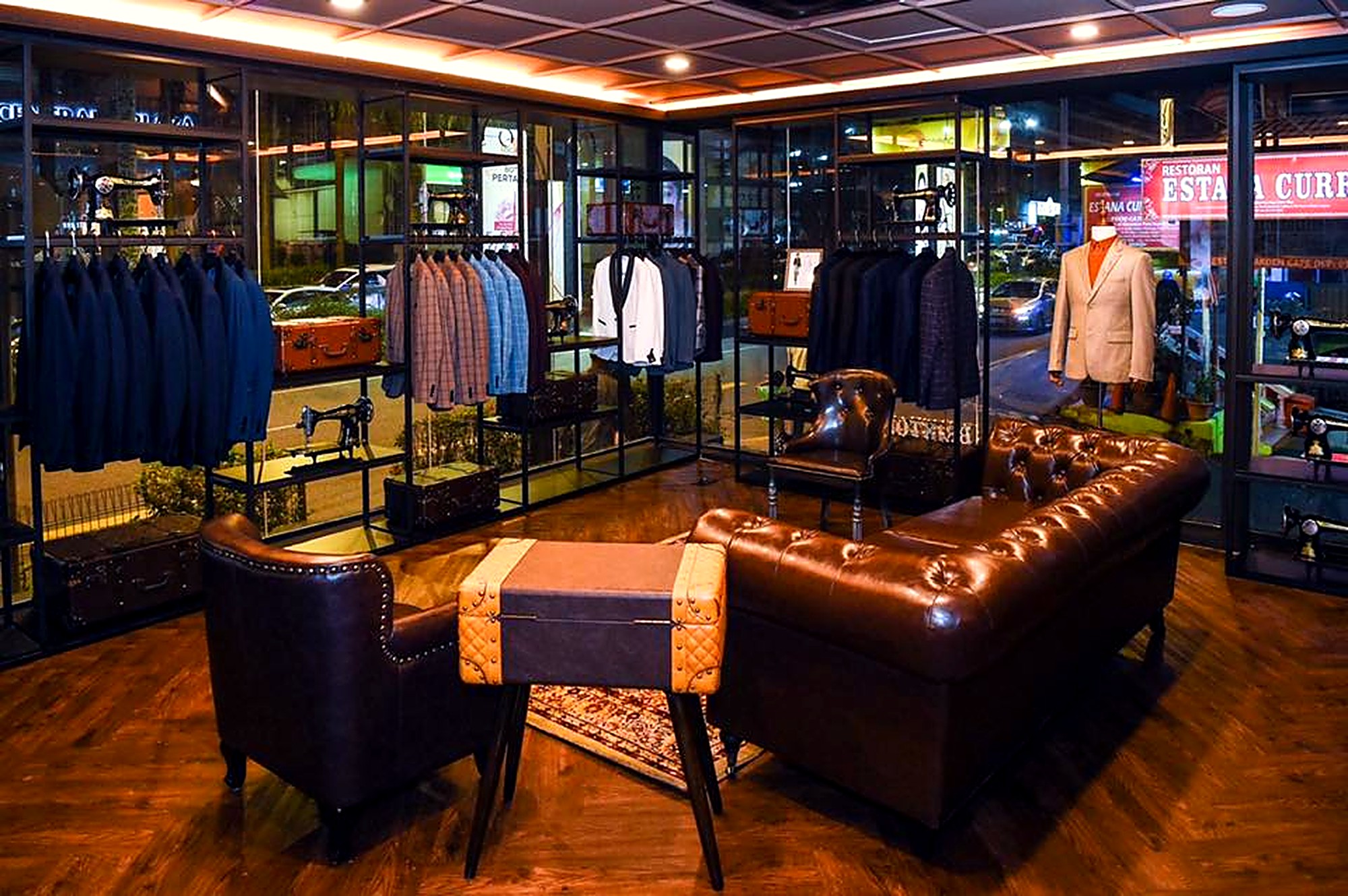 The interior of a tailor shop is shown, with racks of suits and jackets, vintage sewing machines, leather furniture, and display cases